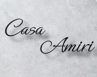 Pode incluir: Um letreiro de parede em metal preto com as palavras Casa Amiri em uma fonte cursiva. As letras são montadas para criar um efeito tridimensional com uma sombra suave projetada na superfície texturizada cinza-claro atrás das letras.