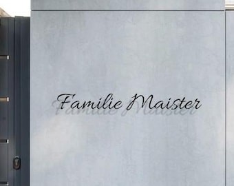 Pode incluir: Uma parede cinza clara com o texto "Familie Maister" em fonte cursiva. A parede é dividida em seções e tem uma aparência texturizada. Uma porta de metal cinza escuro é visível no lado esquerdo.