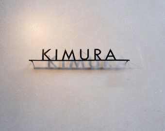 Pode incluir: Uma placa de metal preta que diz "KIMURA" em uma fonte minimalista. A placa está montada em uma parede cinza clara, com as letras projetando uma sombra abaixo. A estética geral é moderna e limpa.