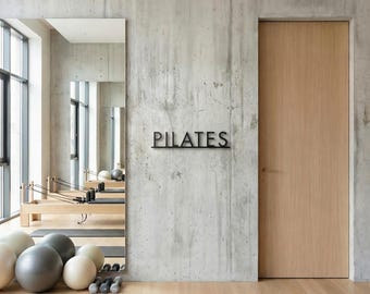 Pode incluir: Interior de um estúdio de Pilates com um grande espelho, equipamentos de exercício e uma porta de madeira. A palavra "PILATES" é exibida em letras pretas na parede de concreto. Bolas de exercício e pesos são visíveis no chão.