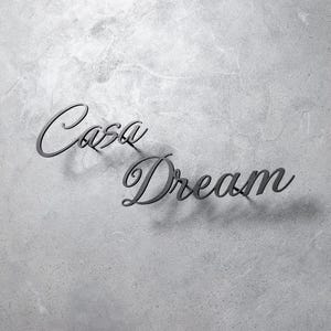 May include: Dark gray metal wall art spelling "Casa Dream" in a cursive font. The letters cast a shadow on the textured, light gray background. The art piece is suitable for home decor.