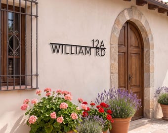 May include: A black metal house sign mounted on a beige wall, featuring the text "WILLIAM RD" and the number "12A" in a clean, sans-serif font. The sign is positioned above several terracotta pots containing pink geraniums and purple lavender.