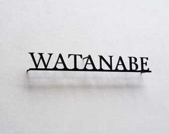 Pode incluir: Uma placa de parede em metal preto com o nome WATANABE numa fonte com serifa. As letras estão ligadas por uma fina barra horizontal na base, criando um design linear e estruturado que se destaca contra a parede clara e lisa.