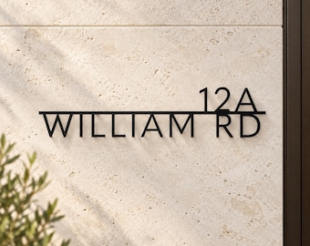 May include: A black metal house sign featuring the text 12A WILLIAM RD in a clean sans-serif font. The sign consists of a horizontal line separating the number 12A from the street name WILLIAM RD, mounted on a light beige stone wall.