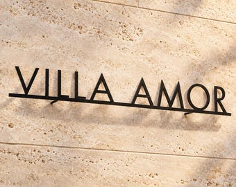 May include: A black metal wall sign featuring the text VILLA AMOR in a clean, sans-serif font. The letters are connected by a horizontal bar at the base, and the entire structure is mounted slightly away from the textured beige stone wall surface.