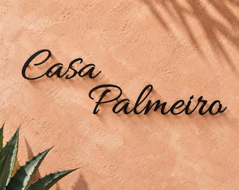 May include: A black metal wall sign featuring the cursive text Casa Palmeiro. The lettering is mounted on a textured, light orange wall surface, with the partial shadow of a palm leaf visible in the upper right corner of the frame.