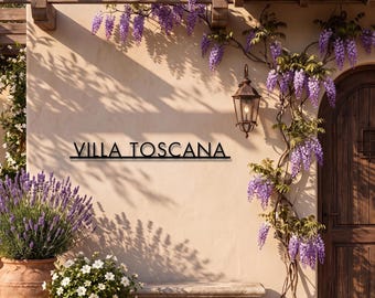 May include: A black metal wall sign featuring the text VILLA TOSCANA in a clean, sans-serif font. The sign is mounted on a light beige stucco wall next to a wooden arched door, a hanging metal lantern, and climbing purple wisteria flowers.