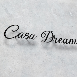 May include: Black metal sign spelling out "Casa Dream" in a cursive font. The sign is mounted on a light gray, textured surface, creating a contrast between the dark letters and the background.