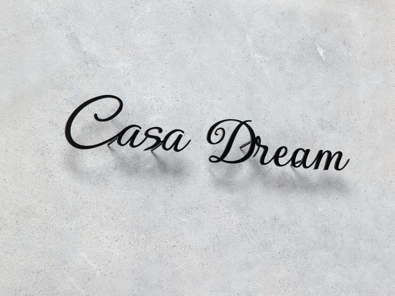 May include: Black metal sign spelling out "Casa Dream" in a cursive font. The sign is mounted on a light gray, textured surface, creating a contrast between the dark letters and the background.