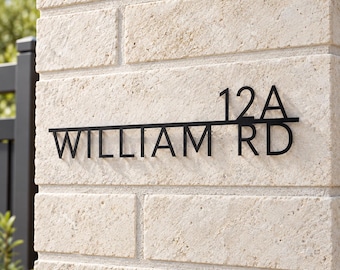 May include: A black metal house number sign mounted on a light beige brick wall. The sign features a horizontal line with the number 12A positioned above the line and the text WILLIAM RD positioned below the line in a clean sans-serif font.