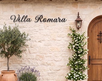 May include: A black metal wall sign with the text Villa Romara in a cursive script font, mounted on a light-colored stone wall next to a wooden arched door, a terracotta planter with an olive tree, and a climbing plant with white flowers.