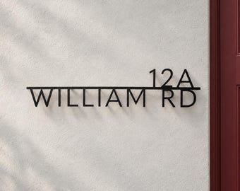 May include: A black metal house sign featuring the text 12A WILLIAM RD in a clean sans-serif font. The sign consists of a horizontal line separating the number 12A on top from the street name WILLIAM RD below, mounted on a textured wall.