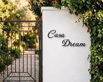 May include: A black metal wall sign featuring the cursive text Casa Dream mounted on a smooth white pillar. The sign is positioned next to a black metal gate and lush green foliage, creating a clear contrast between the dark lettering and the wall.