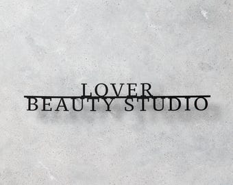 Pode incluir: Uma placa de parede em metal preto com o texto LOVER BEAUTY STUDIO em uma fonte serifada. As letras são conectadas por uma linha horizontal que passa abaixo da palavra LOVER e acima de BEAUTY STUDIO, criando um display de parede unificado.