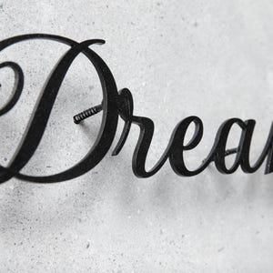 May include: Black metal wall art spelling the word "Dream" in a cursive font. The word is mounted on a textured, light gray surface. The letter "D" has a prominent swirl.