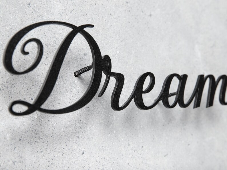 May include: Black metal wall art spelling the word "Dream" in a cursive font. The word is mounted on a textured, light gray surface. The letter "D" has a prominent swirl.
