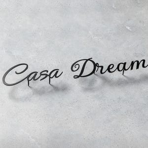 May include: Black metal sign with the words "Casa Dream" in a cursive font. The sign is placed on a light gray, textured surface. The letters are connected and have a shadow.