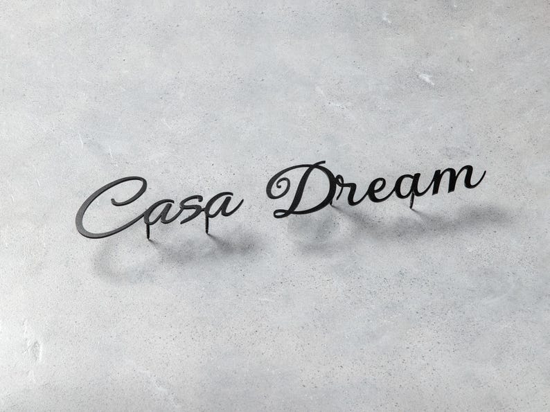 May include: Black metal sign with the words "Casa Dream" in a cursive font. The sign is placed on a light gray, textured surface. The letters are connected and have a shadow.