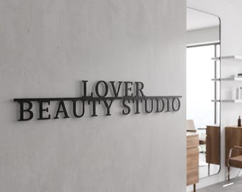 May include: A black metal wall sign featuring the text LOVER BEAUTY STUDIO in a serif font. The word LOVER is positioned above a horizontal line, while the words BEAUTY STUDIO are positioned below the same horizontal line on a wall.