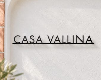 May include: A black metal wall sign featuring the text CASA VALLINA in a clean, sans-serif font. The letters are connected by a thin horizontal line running along the bottom, creating a unified, linear display for mounting on a wall.