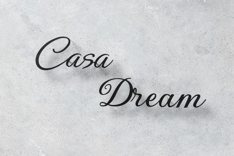 May include: The words "Casa Dream" are displayed in elegant, black cursive script against a speckled, light gray background. The letters cast subtle shadows, enhancing the design's depth.