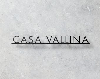 Pode incluir: Placa de metal preta com o texto "CASA VALLINA" em uma fonte minimalista. A placa é montada em um fundo cinza claro texturizado.