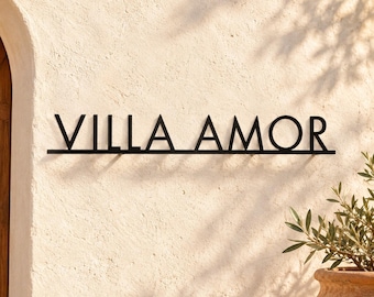 May include: A black metal wall sign featuring the text VILLA AMOR in a clean, sans-serif font. The letters are connected by a horizontal line at the base, creating a singular, unified piece of signage mounted against a textured beige wall.