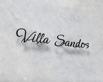 May include: Black cursive text that reads "Villa Sandos" on a light gray, speckled background. The text appears to be a logo or sign, possibly for a business or residence.