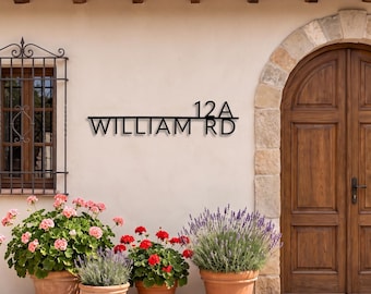 May include: A black metal house sign featuring the text 12A WILLIAM RD in a clean sans-serif font. The sign is mounted on a light-colored exterior wall between a window with a black metal grate and a large wooden arched door.