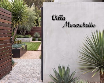 May include: A black metal wall sign featuring the text Villa Moreschetto in a cursive script font. The sign is mounted on a smooth light gray concrete wall, positioned above a garden area with green plants and a stone pathway.