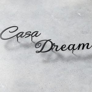 May include: Black metal wall art spelling "Casa Dream" in a cursive font. The words are mounted on small posts, casting shadows on the gray textured surface. Home decor.