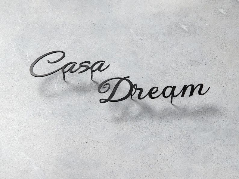 May include: Black metal wall art spelling "Casa Dream" in a cursive font. The words are mounted on small posts, casting shadows on the gray textured surface. Home decor.