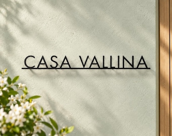 May include: A black metal wall sign featuring the text CASA VALLINA in a clean, sans-serif font. The letters are connected by a thin horizontal line at the base, mounted against a light-colored, textured wall surface.
