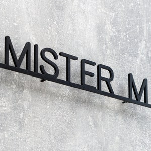 May include: Black metal wall sign that reads "MISTER M" in a sans-serif font. The sign is mounted on a textured gray wall. The letters are connected by a horizontal bar.
