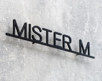 Pode incluir: Placa de parede de metal preto que diz "MISTER M" em uma fonte sem serifa. A placa é montada em uma parede cinza texturizada. As letras são conectadas por uma barra horizontal.