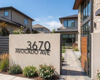 Pode incluir: Uma placa moderna com o número da casa "3670 Avocado Ave" em letras de metal preto. A placa está montada em uma parede de estuque bege, com um jardim paisagístico na frente. A cena é definida sob um céu azul claro.