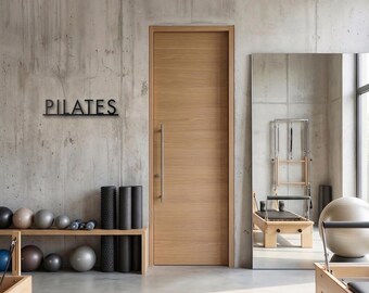 Pode incluir: Interior de um estúdio de Pilates com uma porta de madeira, um espelho grande e equipamentos de exercício. A palavra "PILATES" está na parede. Várias bolas de exercício e rolos de espuma são visíveis nas prateleiras.