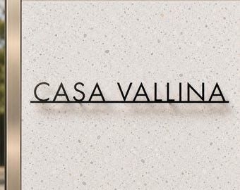May include: A black metal sign featuring the text CASA VALLINA in a clean, sans-serif font. The letters are mounted on a horizontal bar and attached to a light gray, speckled stone wall with a metallic vertical trim on the left side.