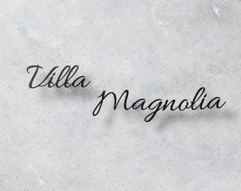 Pode incluir: As palavras "Villa Magnolia" estão escritas em uma fonte cursiva em preto sobre um fundo cinza claro texturizado. O texto tem um ligeiro efeito de sombra, adicionando profundidade ao design.