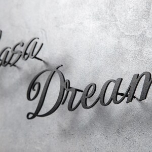 May include: Black metal wall art spelling out "Casey Dream" in a cursive font. The letters are mounted on a textured, gray surface, creating a shadow effect. The artwork is a decorative piece for home decor.
