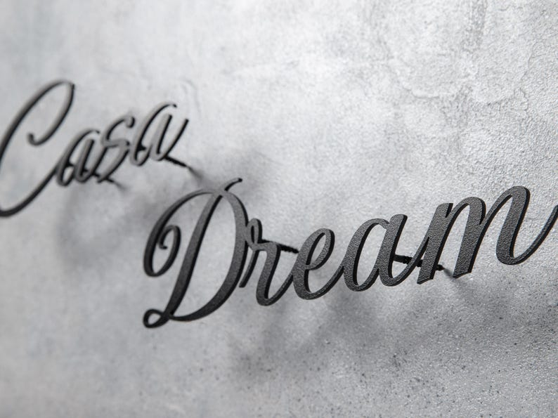 May include: Black metal wall art spelling out "Casey Dream" in a cursive font. The letters are mounted on a textured, gray surface, creating a shadow effect. The artwork is a decorative piece for home decor.
