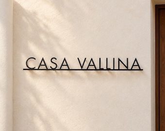 May include: A black metal wall sign featuring the text CASA VALLINA in a clean, sans-serif font. The letters are mounted on a thin horizontal line, creating a dimensional effect against the textured, light-colored wall surface.