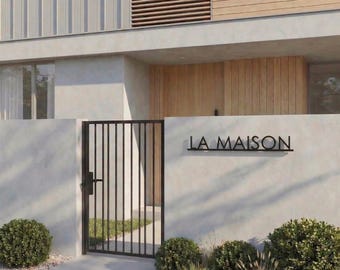 Pode incluir: Vista exterior de uma casa moderna com um portão de metal preto e o texto "LA MAISON" na parede. A casa apresenta paredes de cor clara, detalhes em madeira e paisagismo cuidado com pequenos arbustos e cascalho.