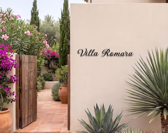 May include: A black metal wall sign featuring the text Villa Romara in a cursive script font, mounted on a smooth beige exterior wall. The sign is positioned above a garden area with green agave plants and terracotta pots.
