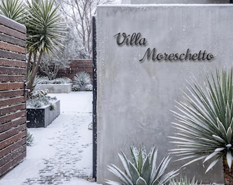 May include: A dark metal sign with the text Villa Moreschetto in a cursive font mounted on a smooth gray concrete wall. The scene includes spiky green plants and a stone path covered in a light dusting of snow during a winter day.