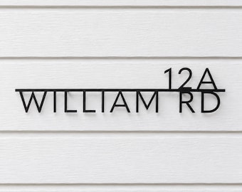 May include: A black metal house sign featuring the text 12A WILLIAM RD. The sign consists of a horizontal line separating the numbers 12A from the text WILLIAM RD, all rendered in a clean, sans-serif font against a white horizontal siding.