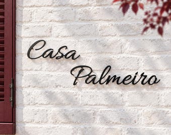 May include: Black metal wall sign featuring the cursive text Casa Palmeiro mounted on a white brick wall. The sign consists of two separate words in a flowing script font, positioned to the right of a dark red shutter.