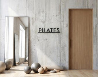 Pode incluir: Interior de um estúdio de Pilates com parede de concreto e piso de madeira. Uma placa de metal preta diz "PILATES". O equipamento de exercício inclui bolas de exercício cinza e de madeira, um tapete de ioga e um espelho de corpo inteiro.