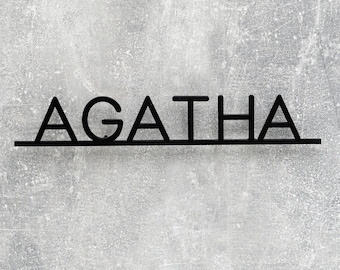 Pode incluir: Uma placa de parede em metal preto com o nome AGATHA em letras maiúsculas. As letras estão ligadas por uma linha horizontal fina ao longo da base, criando um design linear e limpo que se destaca pelo seu acabamento escuro e arrojado.