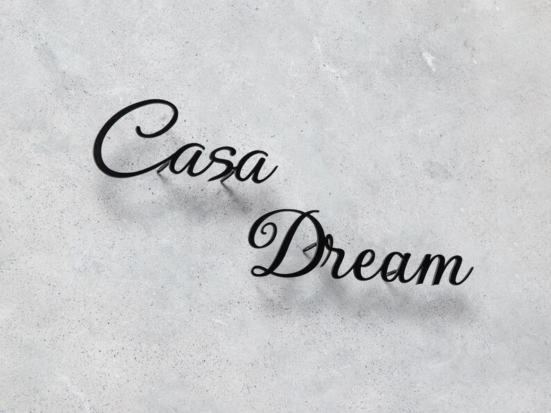 May include: Black metal wall art spelling out "Casa Dream" in a cursive font. The words are mounted on a textured, light gray surface. The letters are slightly raised, creating a subtle shadow.